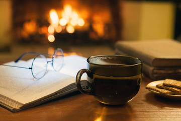An open ancient book, vintage glasses, a cup and biscuits in front of the fireplace. Evening fairy tales. Fantasy.