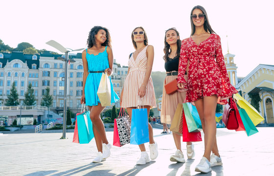 Urban landscape. Full-length photo of four gorgeous feminine women in summer dresses, posing gracefully in different dresses after a shopping spree.