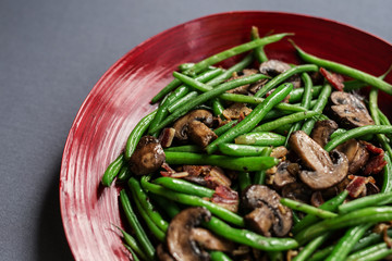 Green beans with mushrooms and bacon. Shown in red bamboo bowl, overhead view on gray background. Copy space.