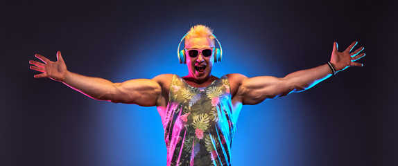 Fashion Muscular DJ man listen to music in colorful neon light with hands up. Handsome blonde...