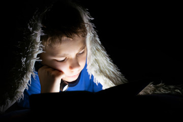 engrossed child reading a book, sitting in a dark room under a blanket and shining a flashlight