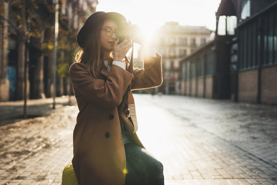 Tourist Girl in hat travels in Barcelona holiday. Photographer in glasses take photo on retro camera. Sunlight flare street in europe city. Traveler hipster shooting architecture, copy space mockup