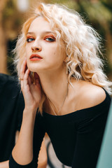 Naklejka premium Exquisite blonde in a black dress with bare shoulders leaning her elbows on a table sits in a cafe