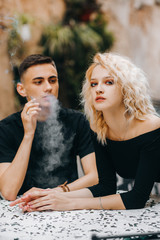 Fototapeta premium Amazing guy with a cigarette in his hand looks at a blond girl in a black dress, standing with her back bent and putting her elbows on the table.