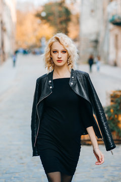 Exquisite blonde in elegant evening black dress and leather jacket walks down the street