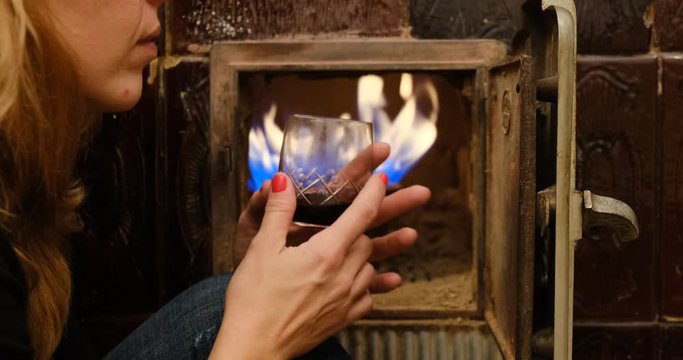 Woman holding red wine glass in front of an old fireplace with gas powered flame inside, during holidays.