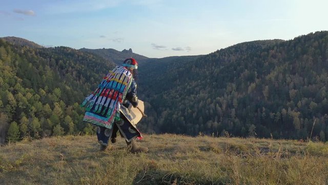 Energetic dance of an old shaman with a tambourine on the background of autumn mountain landscape.
