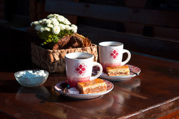 Morning breakfast: hot tea in two white cups and cakes on white flower bouquet background at wooden table