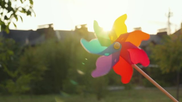 Plastic rainbow windmill rotates on the background of blurry houses and sunset. Suburbs Concept