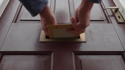 Slow motion close POV shot, from ground level, of a man's hands posting a flat package / letter through the brass letterbox of a house with traditional front door.