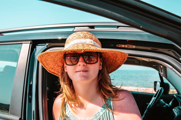 Naklejka premium A woman in hats and sun glasses sits in a dark car. Car parked close to the beach and a view of the sea. The young girl travels during the holidays and stopped to rest.