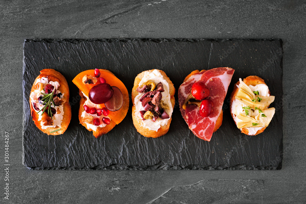 Wall mural Mixed crostini appetizers with a variety of toppings. Top view on a dark slate background. Party food concept.