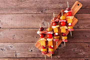 Skewer appetizer board with meat, cheeses and pickles. Above view over a rustic wood background...
