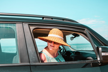 Young woman in a straw hat and sunglasses. Dark soul car on the beach. View of beautiful sea and...