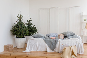 Fototapeta na wymiar Stylish white bedroom, minimalism, Scandinavian style. White linens, knitted gray plaid, pillows. Wooden floor. There are Christmas trees with garlands of lights by the bed. Pink white meringue. Card