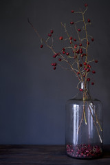Large glass bottle with branches of dry red rosehip on wood table, selective focus