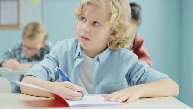 In Elementary School Class: Portrait of a Brilliant and Cute Caucasian Boy Writing in Exercise Notebook. Junior Classroom with Diverse Group of Bright Children Working Diligently, Learning New Stuff