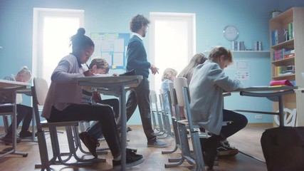 In Elementary School Class: Enthusiastic Teacher walks between Rows of Diverse Children, Explains Lesson. Group of Smart Multiethnic Kids Learning New Stuff. Side View Tracking Shot in Slow Motion