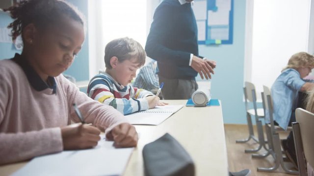 In Elementary School Class: Enthusiastic Teacher walks between Rows of Bright Diverse Children, Dictates Lesson. Group of Smart Multiethnic Kids Writing in Exercise Notebooks. Slow Motion 