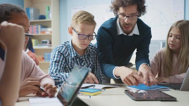 Elementary School Classroom: Enthusiastic Teacher Holding Tablet Computer Explains Lesson to Brilliant Young Children. Kids Learning Programming Languages, Internet Safety and Software Design