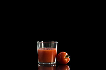 a glass cup with tomato juice stands on a reflective surface on a dark background next to it lies a red round tomato.