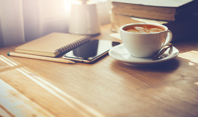 Closeup latte art coffee in white cup with smartphone and notebook on wooden table with sunlight through the window. Vintage color tone