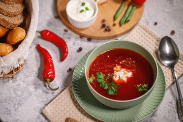 Tasty and hearty dinner. A plate with borsch on the table, next to the board are vegetables, parsley, dill, green onions, garlic, chili peppers, cherry tomatoes, and bread.
