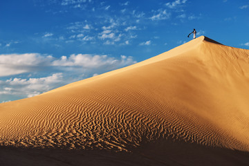 A traditional dressed moroccan man stands on a sand dune against a cloudy blue sky.