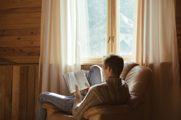 Young man in warm sweater reading by the window inside cozy log cabin - obrazy, fototapety, plakaty