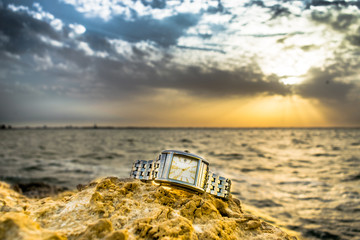an exotic stainless big dial silver watch kept on brownish rocks at the beach and greenish sea water in the background and clouds on the sky