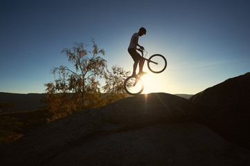 Silhouette of professional cyclist standing on back wheel on trial bicycle. Sportsman rider balancing on the edge of big boulder on the top of mountain at sunset. Concept of extreme sport