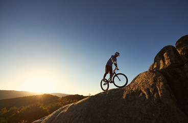 Silhouette of professional bicyclist balancing on trial bicycle on top of big boulder, male rider making acrobatic stunt on summer evening, blue sky and sunset on background. Concept of extreme sport