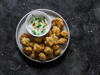 Baked crispy parmesan bread crumbs cauliflower bites with yogurt sauce on dark background, top view