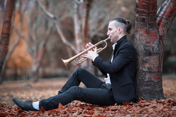 young slender man in black suit plays golden jazz trumpet sitting near brown tree with many fallen...