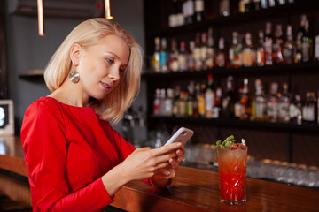 Young beautiful woman in red dress using her smart phone at the bar. Woman typing message on her phone while having cocktail
