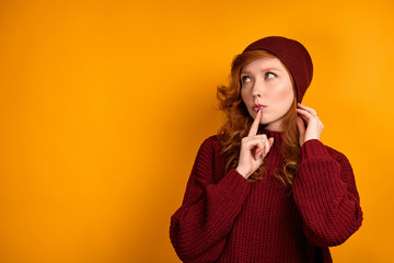 Red-haired girl in a burgundy sweater stands on an orange background and with a finger to lips, straightens her hat and looks up