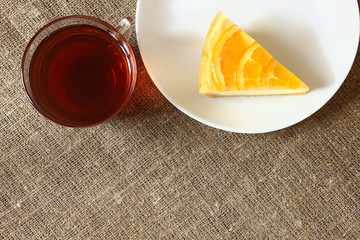 Cheesecake with oranges on a white plate, glass mug with tea on burlap tablecloth. Top view.