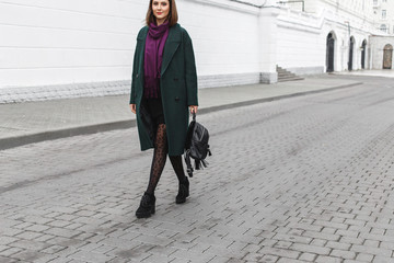 Beautiful young stylish brunette woman wearing midi green coat, black heeled boots, backpack, leopard print tights, purple scarf walking through the city streets. Trendy casual outfit. Street fashion.