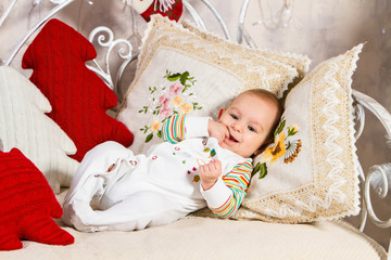 baby with fingers of his right hand in his mouth lying on a sofa among pillows and toys on New Year's theme