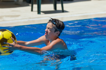 Handsome teenager boy wearing black goggles playing with ball in swimming pool outdoors in a sunny summer day