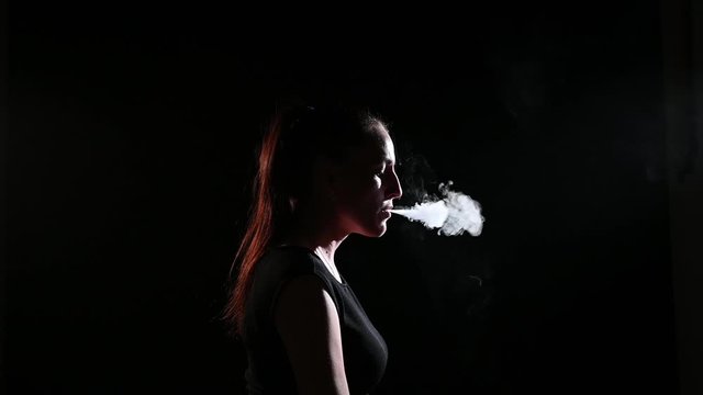 The brunette smokes an electronic cigarette on a black background, releases thick smoke from her mouth. Portrait of a woman smoking a vape, hovering.