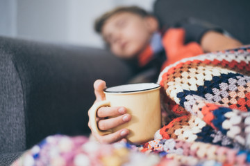 Sick boy sleeping with woolen blanket, hot water bottle and a mug. Sad teen with the flu rests alone at home in a cold winter day. Child with seasonal infections and fever Soft focus on the cup.