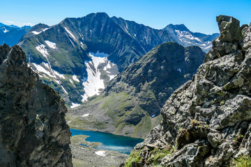 A distance view on a clear, navy blue lake hiding between tall mountain peaks. Some of the slopes are still covered with snow. In the back here is another mountain range. Clear and bright day.