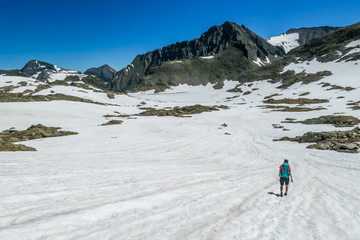 A young woman with a big backpack hikes through a snowy plateau in high mountains. Girl is wearing shorts.In the back there is a sharp mountain peak. Schladming Alps in Austria. Outdoor activities. 