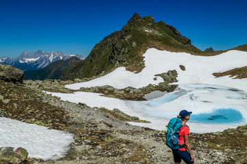 Obraz premium Young woman enjoying the view a frozen alpine lake. The ice shines in many different shades of blue. Lake is surrounded by tall mountains. No snow on the peaks Spring slowly coming to high altitudes