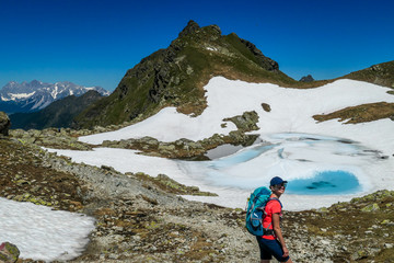 Fototapeta premium Young woman enjoying the view a frozen alpine lake. The ice shines in many different shades of blue. Lake is surrounded by tall mountains. No snow on the peaks Spring slowly coming to high altitudes