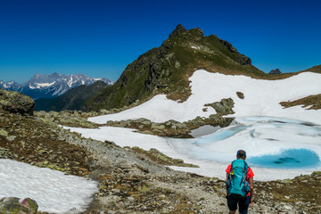 Obraz premium Young woman enjoying the view a frozen alpine lake. The ice shines in many different shades of blue. Lake is surrounded by tall mountains. No snow on the peaks Spring slowly coming to high altitudes