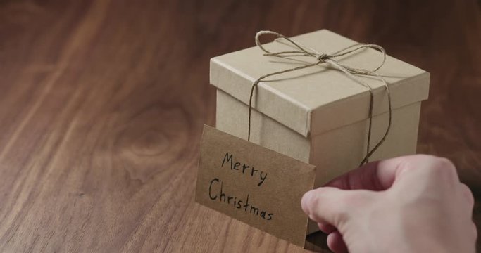 Slow motion man hand put merry christmas card next to brown paper gift box on walnut table