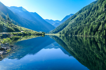 Fototapeta na wymiar A calm alpine lake. The lake is surrounded with tall mountains. The surface of the lake is calm, it reflects the mountains and sky. Clear and sunny day. Schladming region, Austria