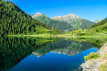 Naklejka na ściany i meble A calm alpine lake. The lake is surrounded with tall mountains. The surface of the lake is calm, it reflects the mountains and sky. Clear and sunny day. Schladming region, Austria